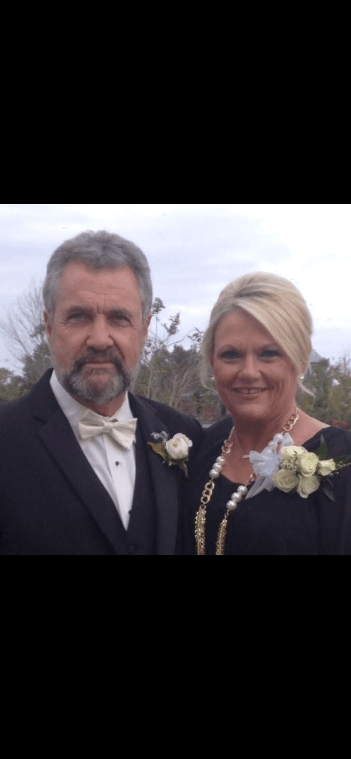 Smiling blonde woman and bearded man in formal attire with white rose corsages and boutonnieres.