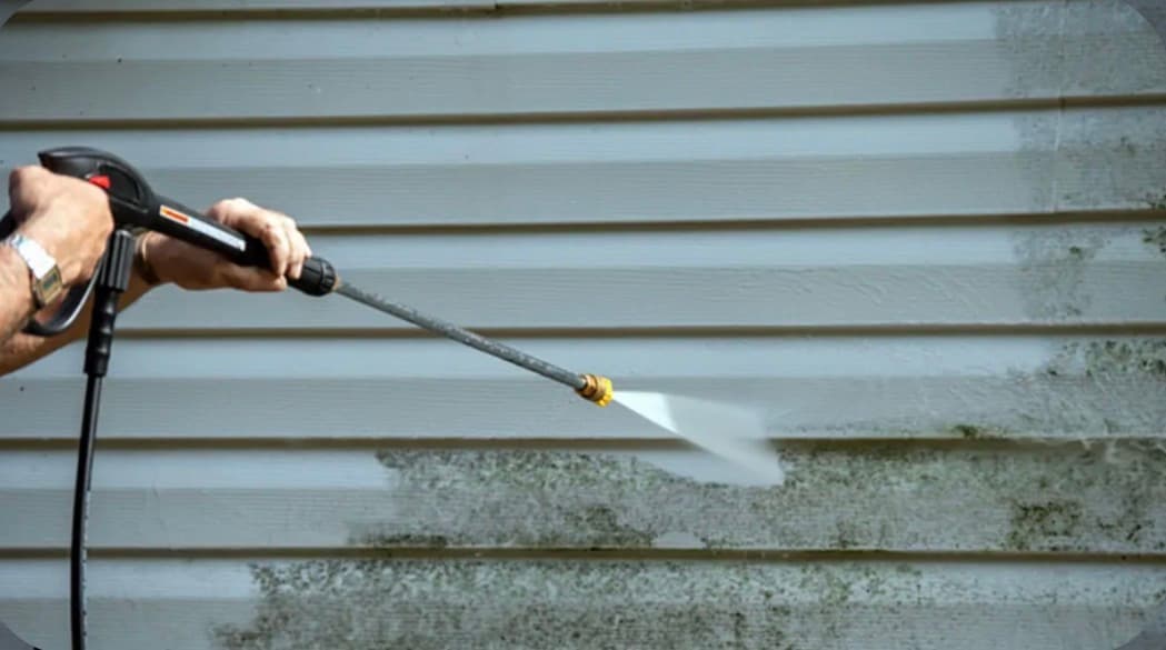 Hands using a pressure washer to remove dirt and mold from white horizontal siding.