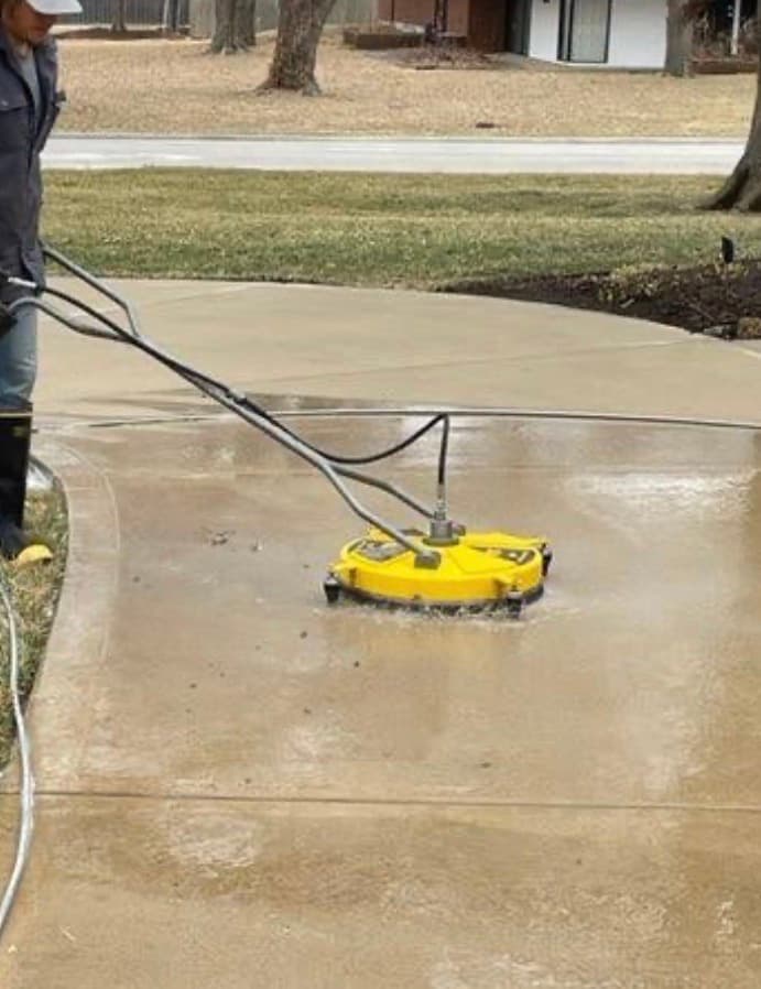 A person uses a yellow circular surface cleaner to pressure wash a wet concrete driveway.