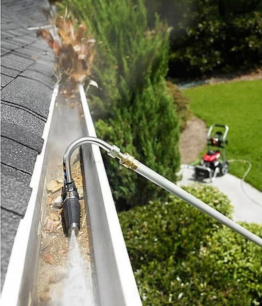 Curved pressure washer wand attachment clearing leaves and debris from a residential roof gutter.