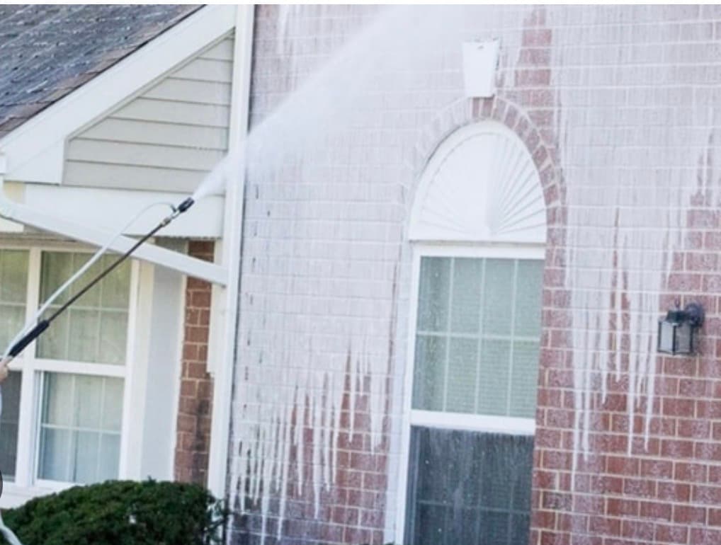 Pressure washer spraying white foam onto a red brick house wall near an arched window.